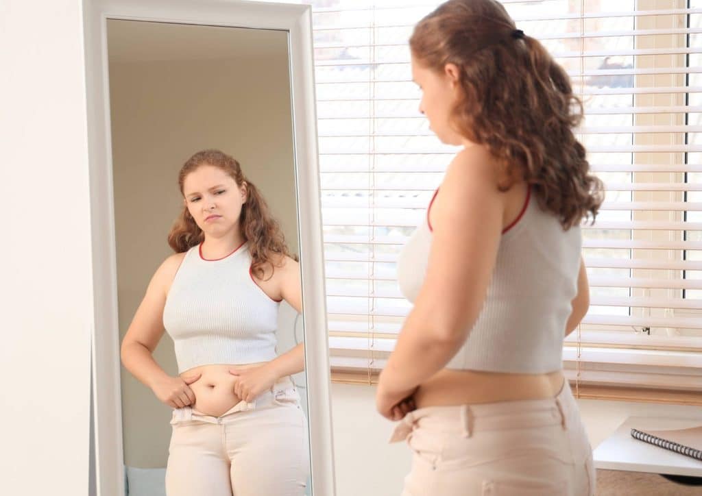 Postpartum Weight Loss: A Gentle, Realistic Guide 8 A woman stands in front of a mirror, gazing at her reflection with a thoughtful expression.
