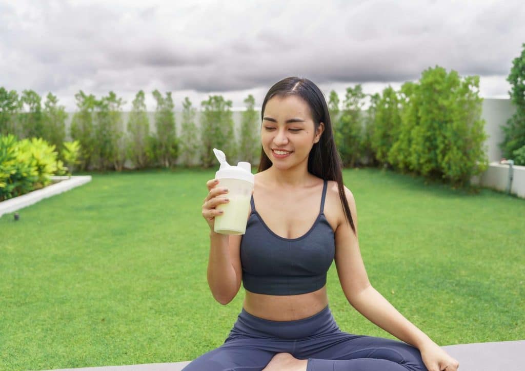 Is Whey Protein Good for Weight Loss? 13 Asian woman in a sports bra holds a smoothie, smiling, showcasing a healthy lifestyle and fitness enthusiasm.