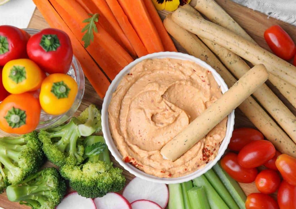 High Fibre Diet for Weight Loss: Why Fibre Keeps You Full 10 A wooden cutting board displaying an assortment of fresh vegetables, a bowl of dip, and several breadsticks.